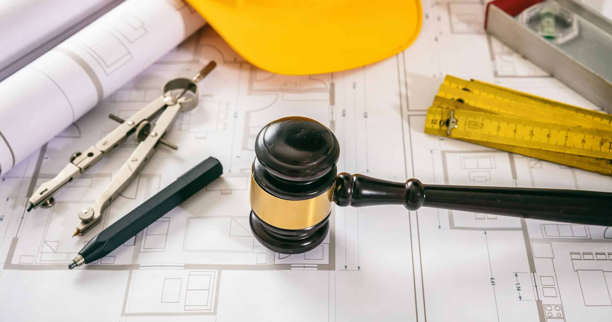 A judge’s gavel on building blueprints beside a hard hat, a folding ruler, and drafting tools