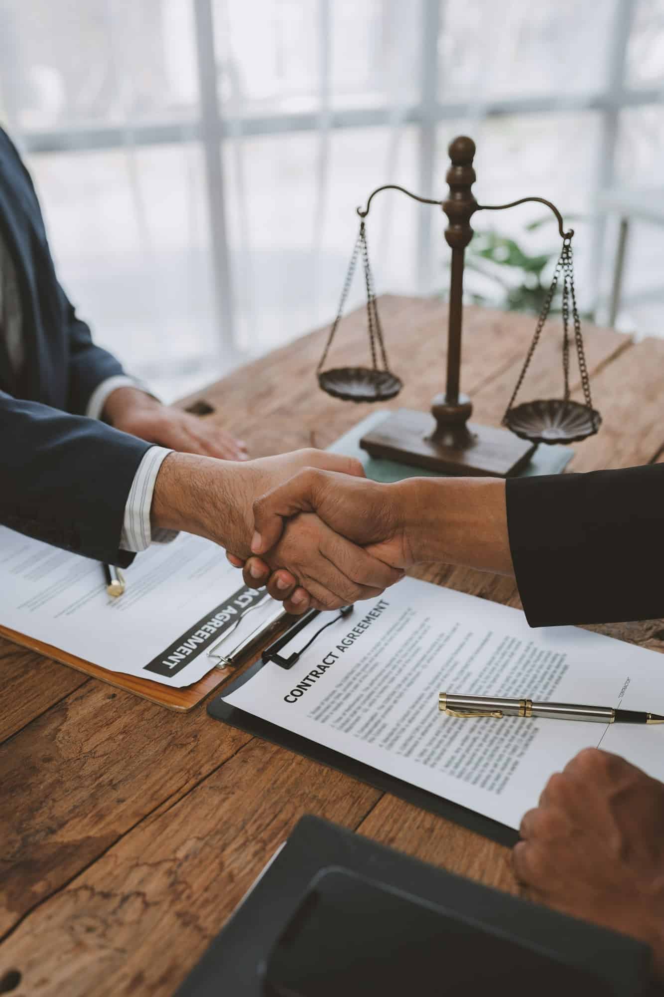 An attorney shakes hands with a client across a desk with a signed contract and the scales of justice