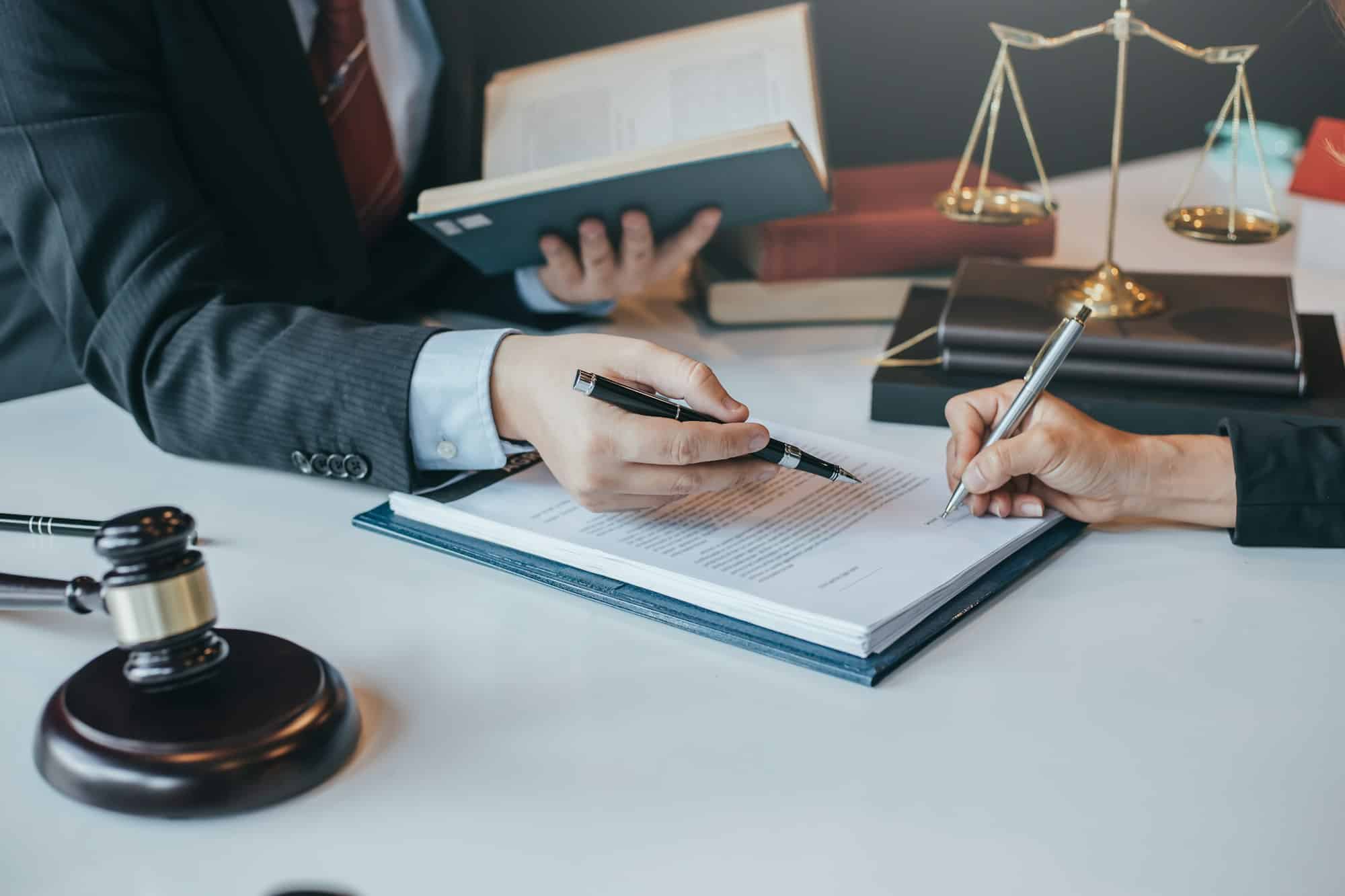 A lawyer and a client sign a legal agreement beside a gavel and the scales of justice